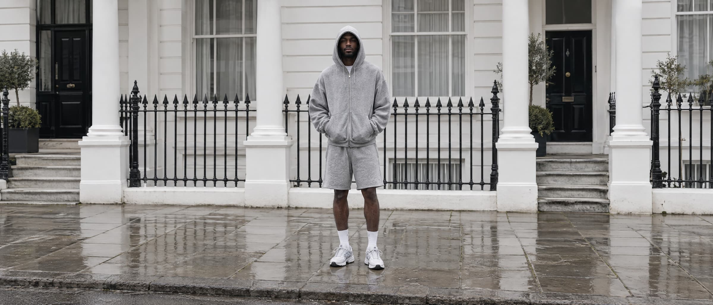 A model in a grey marl zip hoodie and matching shorts standing on a wet pavement in front of a white stucco London terrace with black iron railings.