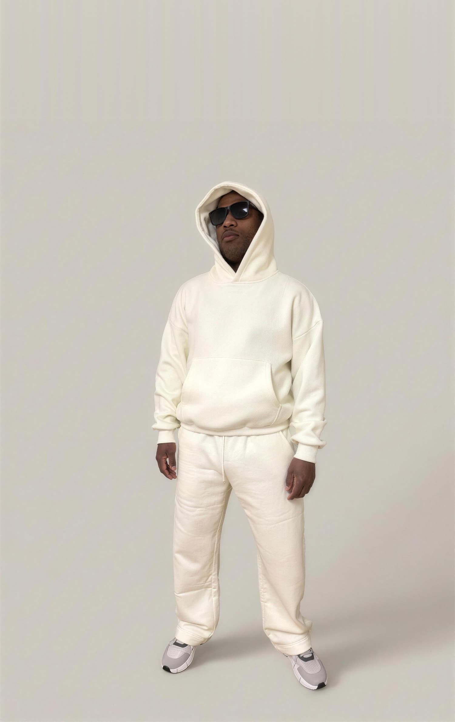 A model wearing the Kensington Pullover Hoodie and Kensington Trouser in cream, hood up, against a plain studio backdrop